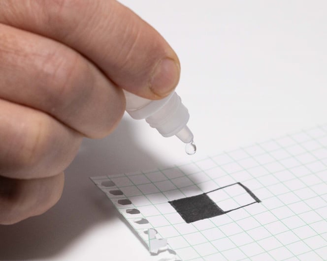 A drop of water coming out of a eye dropper onto a piece of paper with one black graphite box and one white paper box drawn.