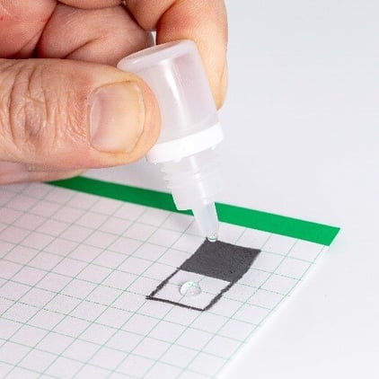 A drop of water coming out of a eye dropper onto a piece of paper with one black graphite box and one white paper box drawn.