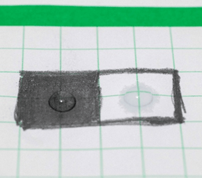 At 15 seconds, a drop of water sitting on top of a black graphite box and a second smaller drop of water sitting on top of a white paper box with a ring of water around it that has soaked into the paper.