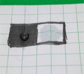 At 15 seconds, a drop of water sitting on top of a black graphite box and a second much smaller drop of water sitting on top of a white paper box with a very large ring of water around it that has soaked into the paper.
