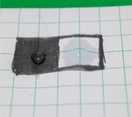 At 15 seconds, a drop of water sitting on top of a black graphite box and a second much smaller drop of water sitting on top of a white paper box with a very large ring of water around it that has soaked into the paper.