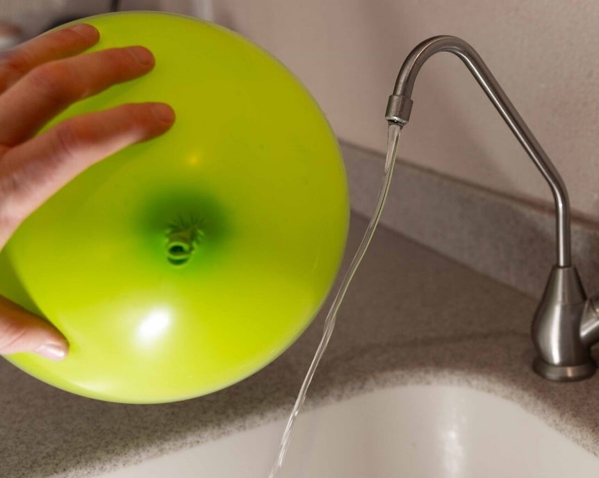 A hand holding a balloon by a small stream of water from a faucet. The water does not fall straight from the faucet, but is angled toward the ballon.